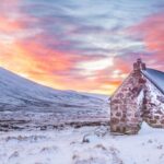 Slopes - Brown House Surrounded by Snow Covered Field Near Snow Covered Mountain Under Yellow Blue and Orange Sunset